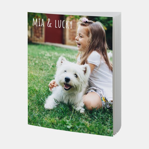 Das echte Fotobuch A4 Hoch Softcover zeigt ein lachendes Mädchen mit weißem Hund auf einer grünen Wiese.