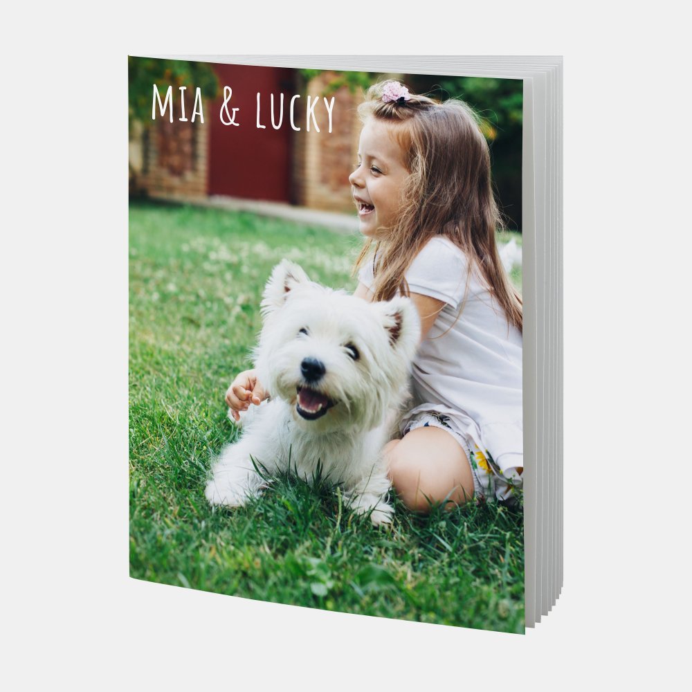 Das echte Fotobuch A4 Hoch Softcover zeigt ein lachendes Mädchen mit weißem Hund auf einer grünen Wiese.