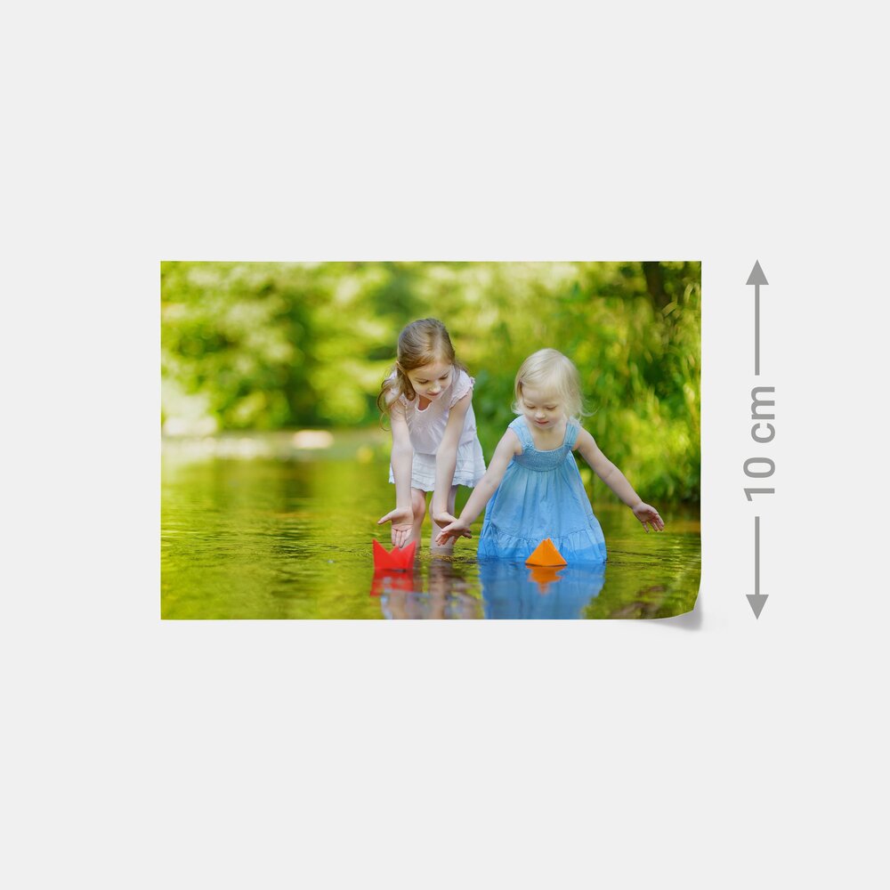 Das Bild zeigt zwei Mädchen, die im Sommer an einem Bach Papierboote treiben lassen – ideal für Fotoabzüge in 10 cm Höhe.