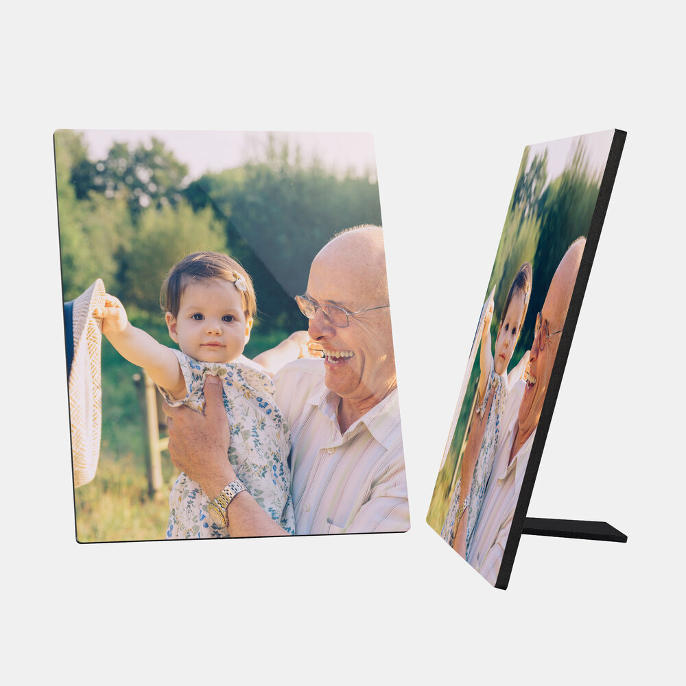 Ein quadratischer Fotoaufsteller zeigt ein schönes Bild von einem älteren Mann, der ein kleines Kind im Arm hält. Der Mann trägt eine helle, gestreifte Bluse und eine Brille und lächelt herzlich. 